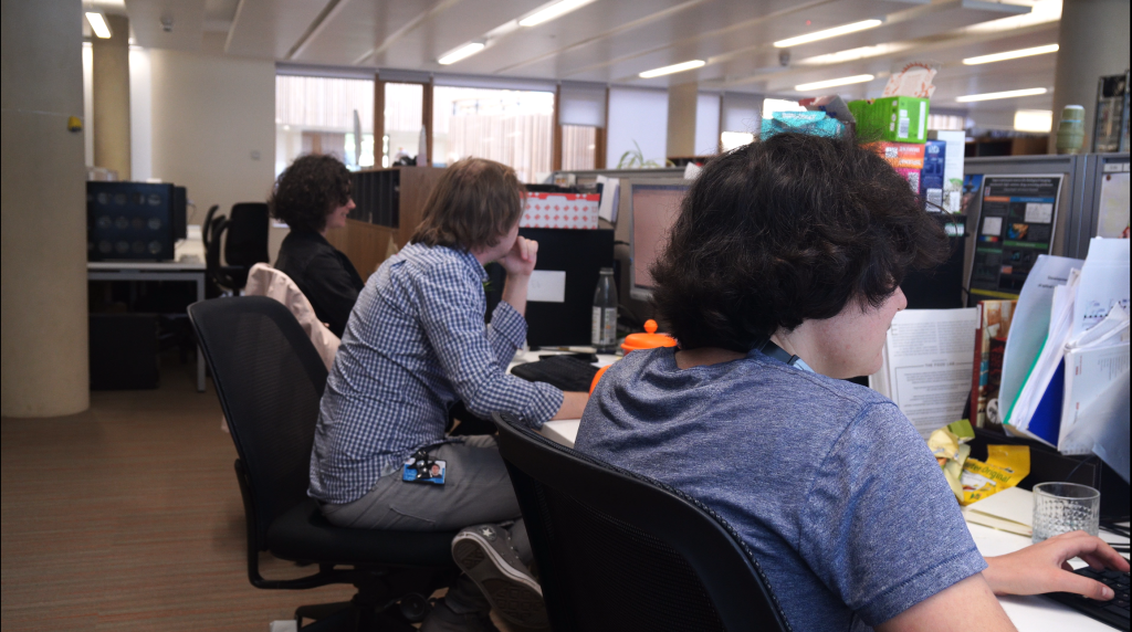 Three people sit at a long desk in an office. Each works at an individual computer