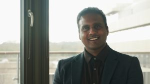 Akshay Deshmukh smiles at the camera in dark suit with West Cambridge in background.