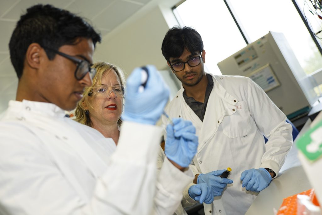 A scientist in a lab coat uses a pipette while supervised by two other scientists.