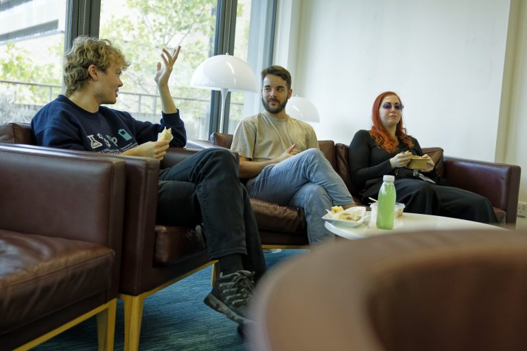 Three people in casual clothing sitting on maroon leather couches on bright day looking relaxed and eating.