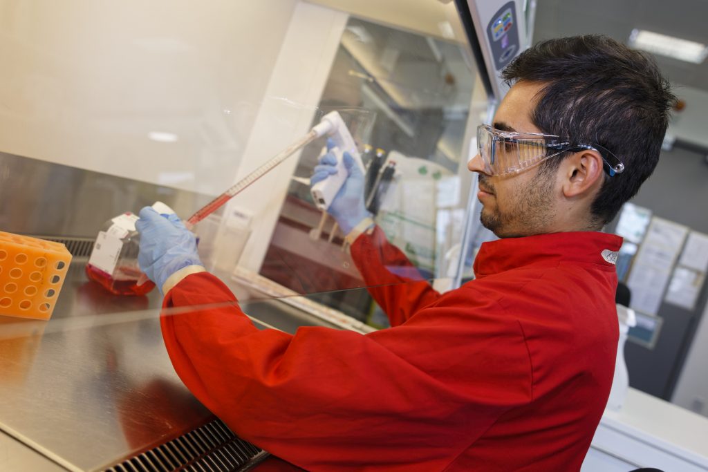 Man in red lab coat and safety glasses in blue gloves pipettes a liquid in a fume hood.