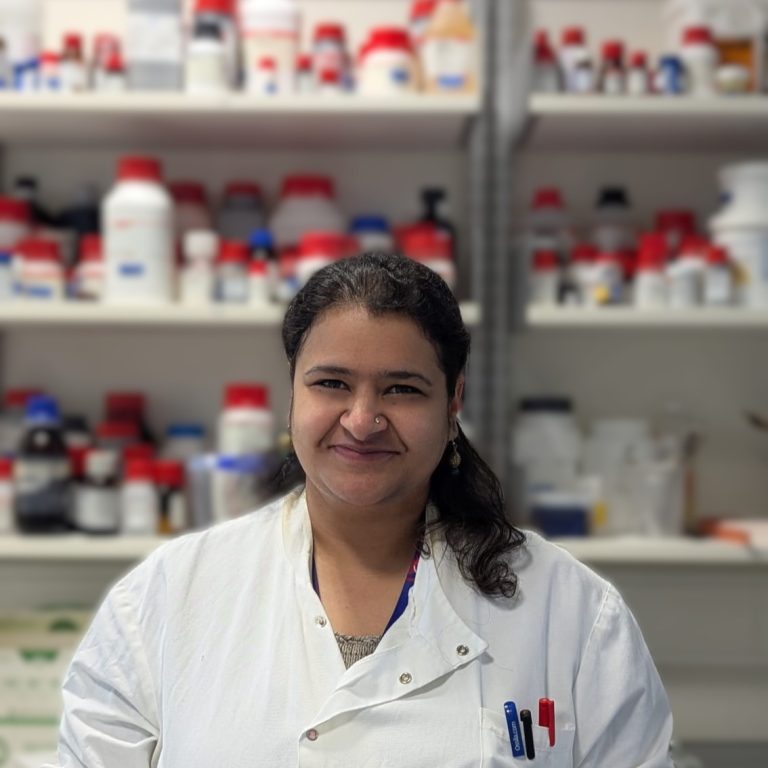A portrait of a woman in a lab coat smiling at the camera. A lab environment forms the background