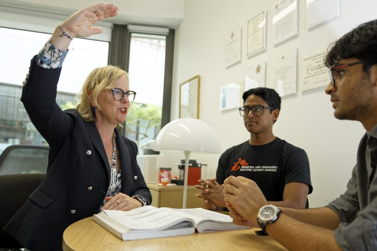 Woman holds hand up to indicate height as she talks to two young men in an office setting.