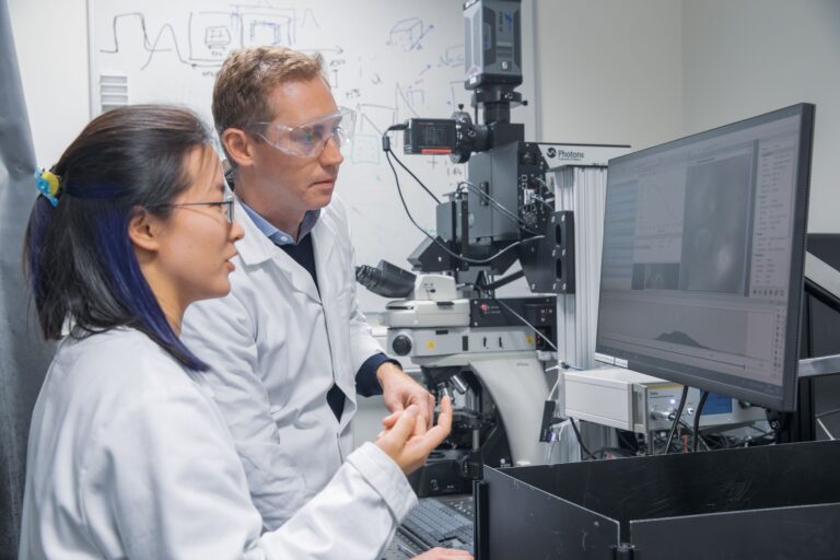 Prof Sam Stranks with female group member in lab coats in lab setting with computer and laser in front of them.