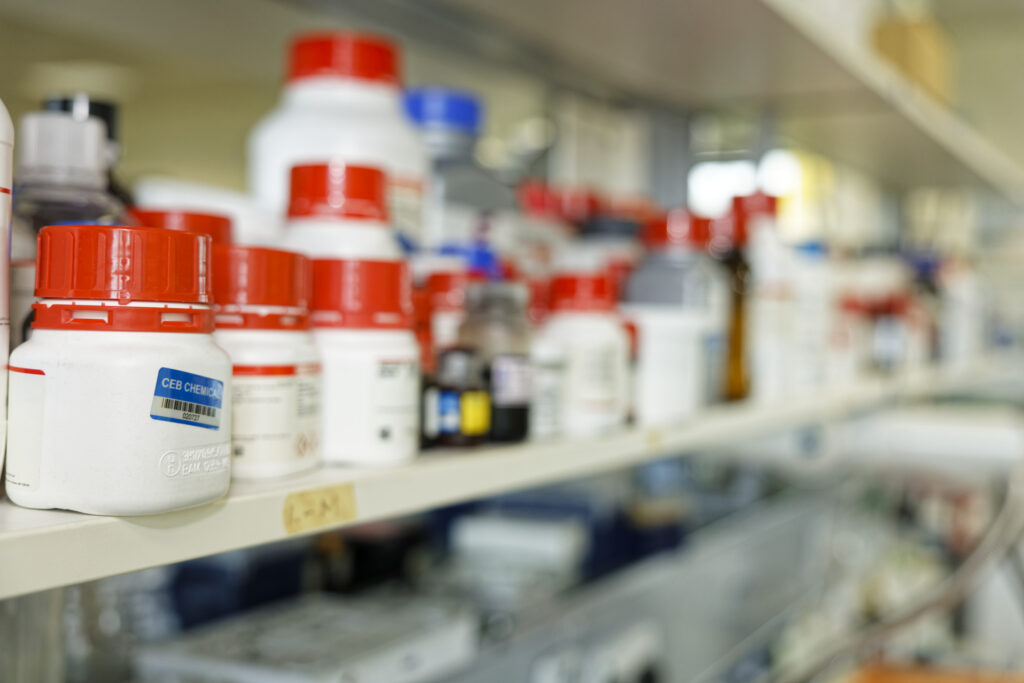 Image of white pill bottles with red tops on a shelf in lab setting.