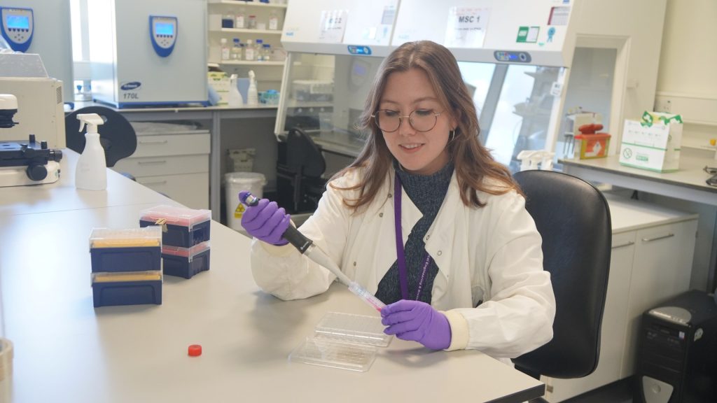 Female scientist in white lab coat pipettes in large lab setting.
