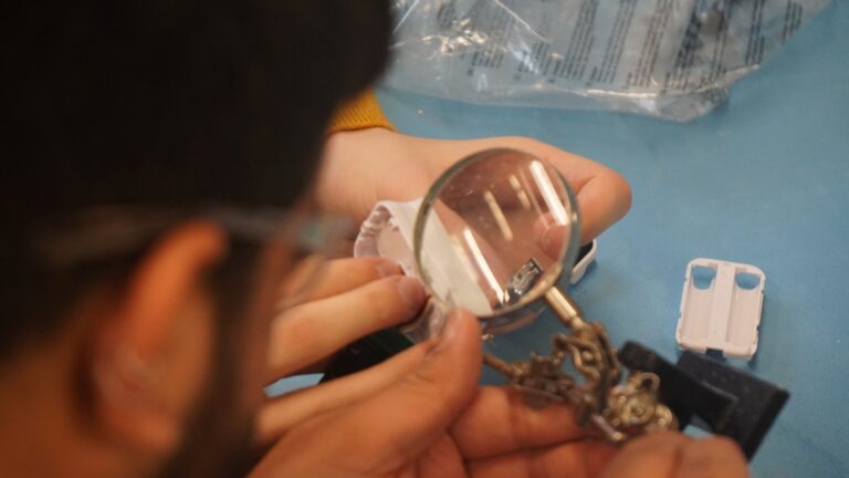 Two people work up close with electrical item under magnifying glass. Man's face up close out of focus on left of image.