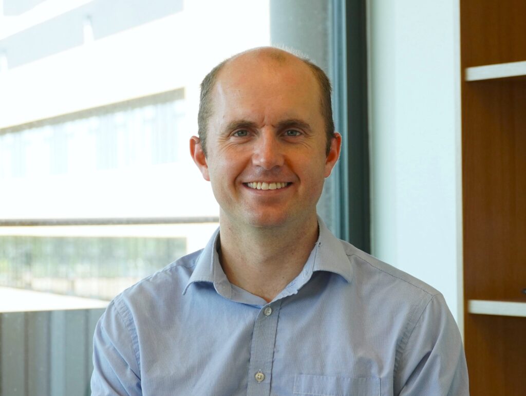 Dr George Gordon in light blue, open button shirt smiling at the camera in office setting.