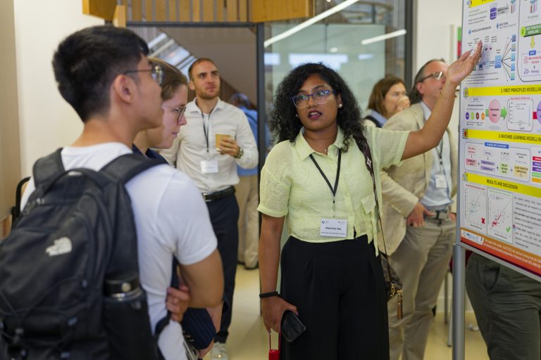 Woman in yellow top explains scientific poster to two people in busy academic setting.
