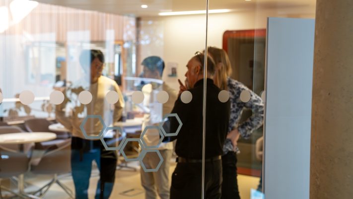 Group of men stand together in open space inside CEB building, looking through glass.