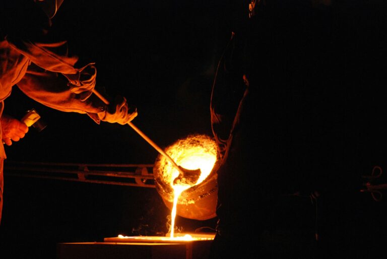 A person is helping to direct molten metal into a mold from a burning hot pot of liquid metal.